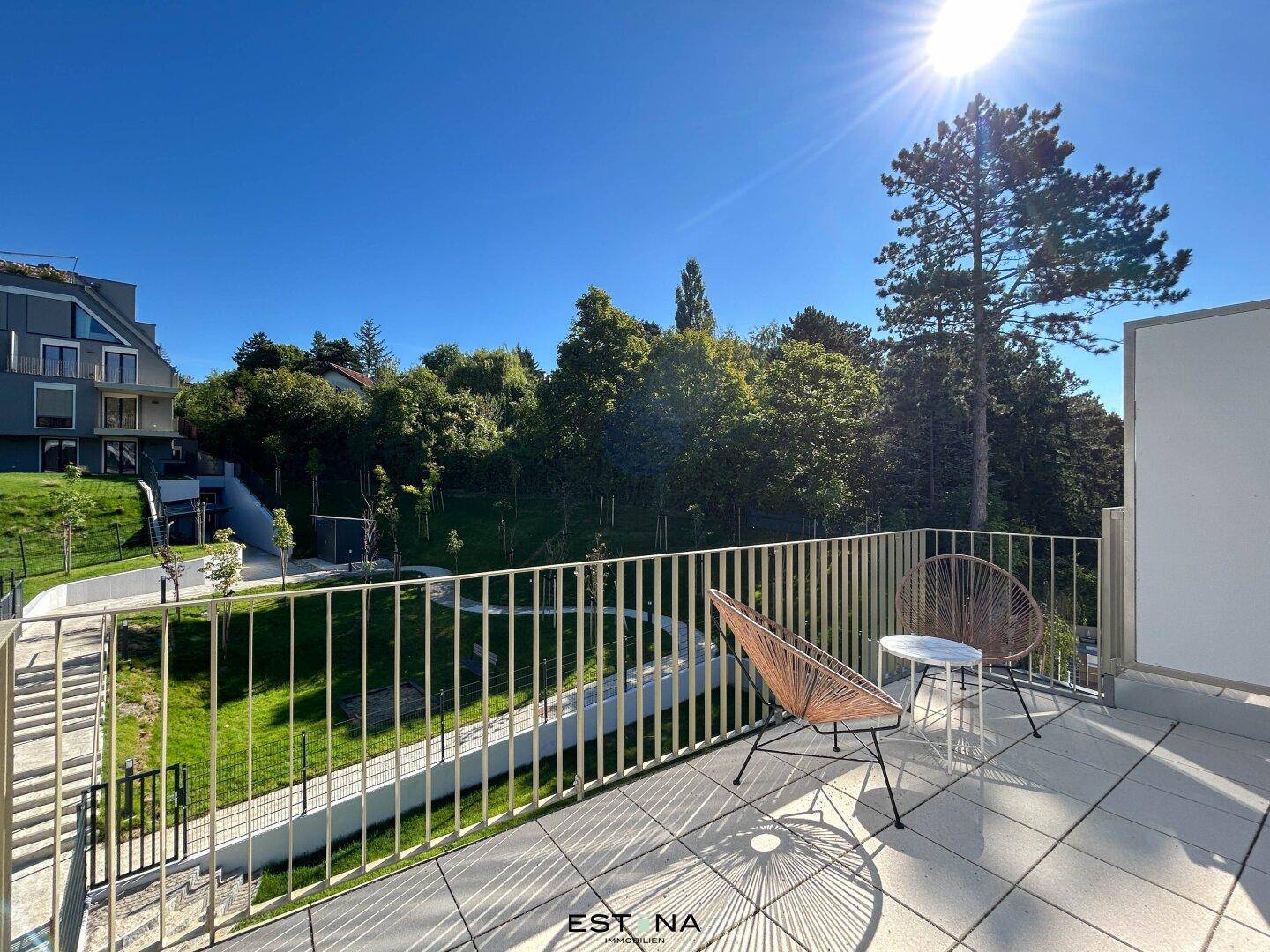 Sonniger Balkon mit Sitzgelegenheiten und Blick auf die grüne Umgebung und weitere Gebäude.