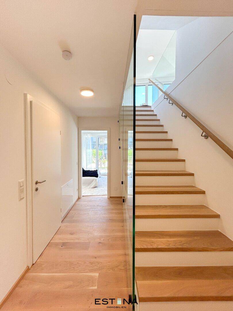 Heller Flur mit Holztreppe, Glasgeländer und Zugang zu weiteren Räumen.