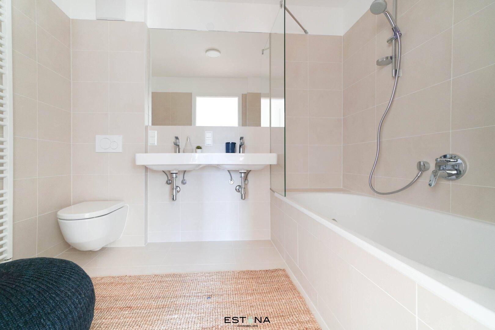 Modernes Badezimmer mit Badewanne, Doppelwaschbecken und Wand-WC, hell gefliest.