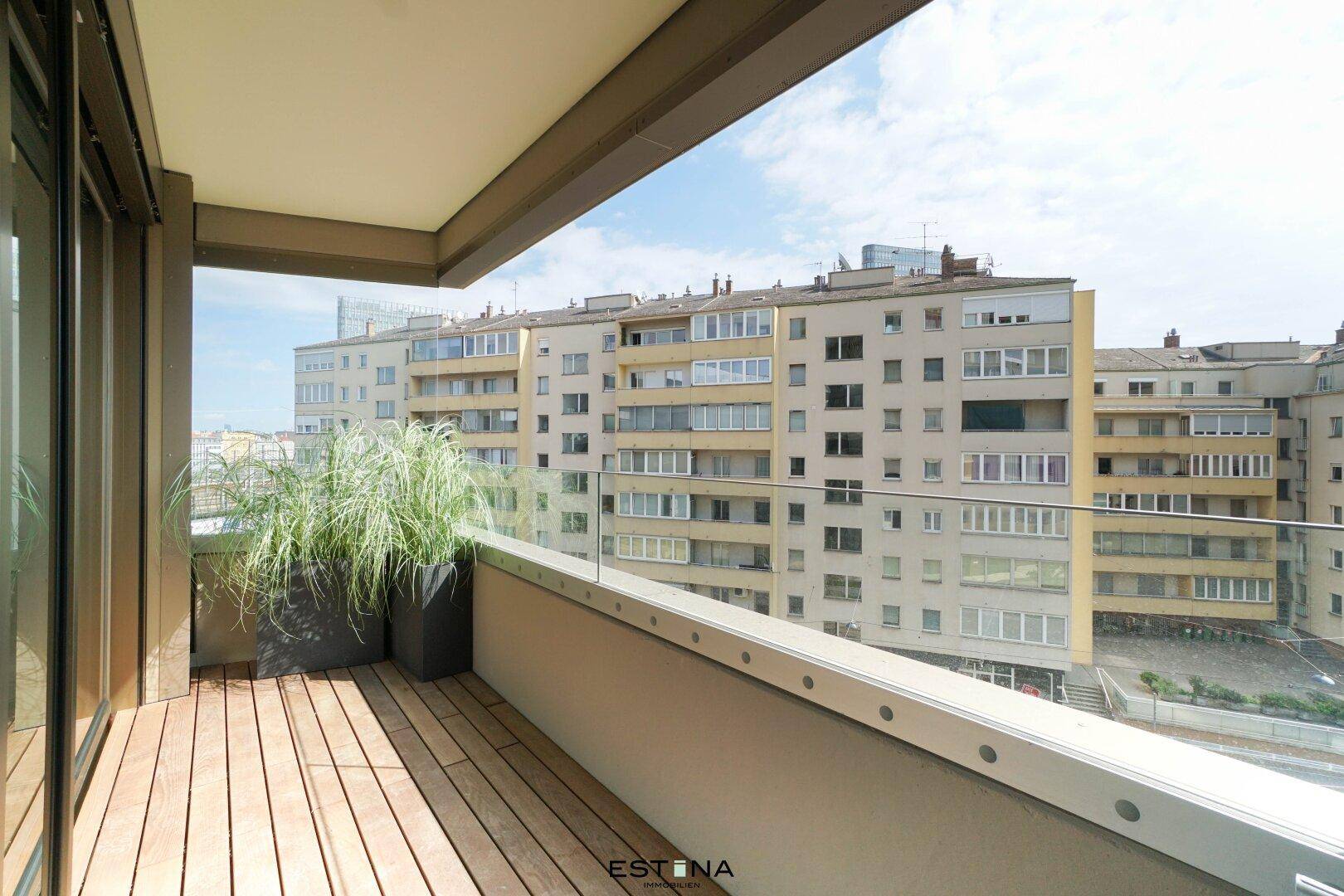 Geräumiger Balkon mit Holzboden und Glasgeländer, bietet Blick auf umliegende Gebäude.