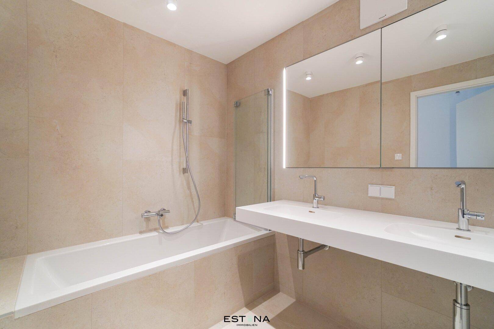 Modernes Badezimmer mit beigefarbenen Fliesen, Badewanne und Doppelwaschbecken mit Spiegelschrank.