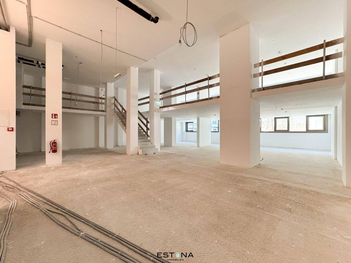 Geräumiger Innenraum im Rohbau mit sichtbaren Säulen und Treppen zu einer Galerie.