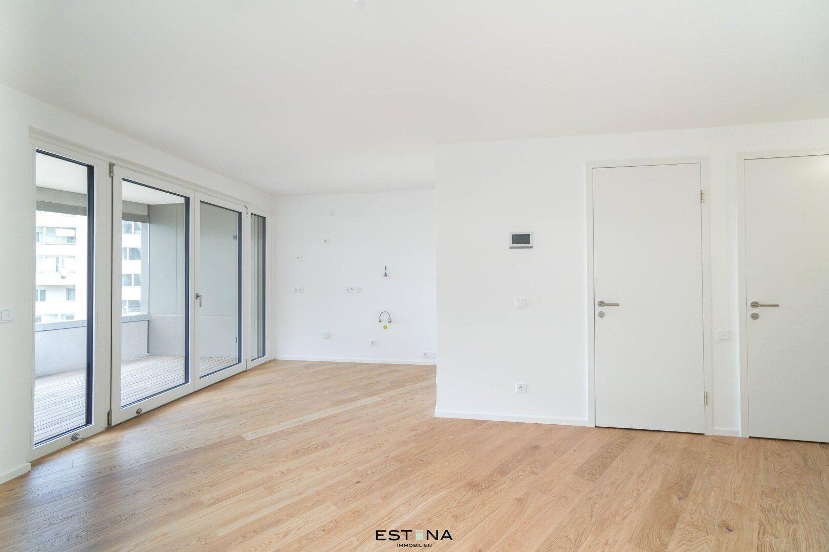Leeres Wohnzimmer mit Holzboden und Zugang zu einem Balkon durch große Glastüren.