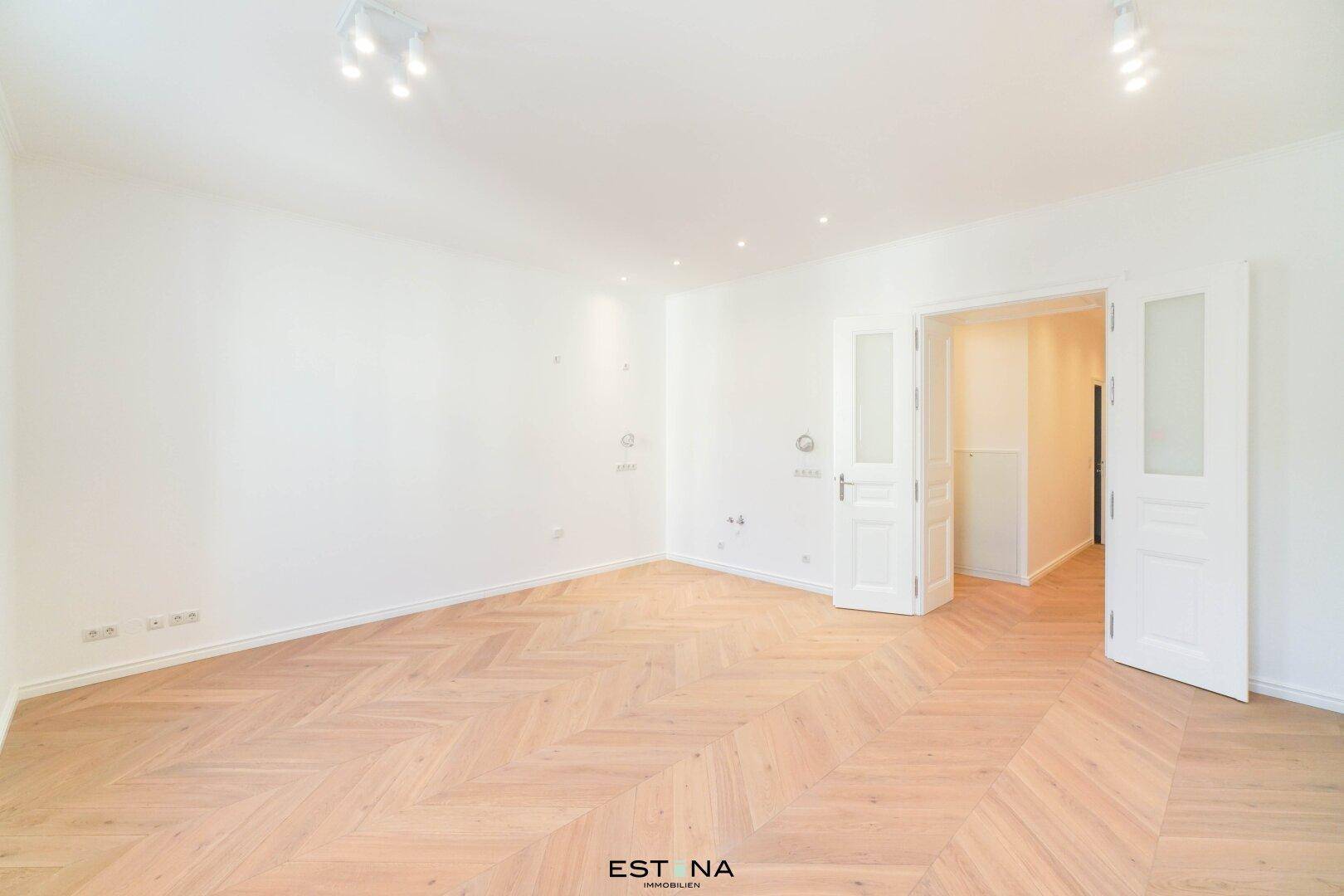 Geräumiges Zimmer mit hellem Fischgrätparkett und doppelten Flügeltüren, ideal für offenes Wohnen.