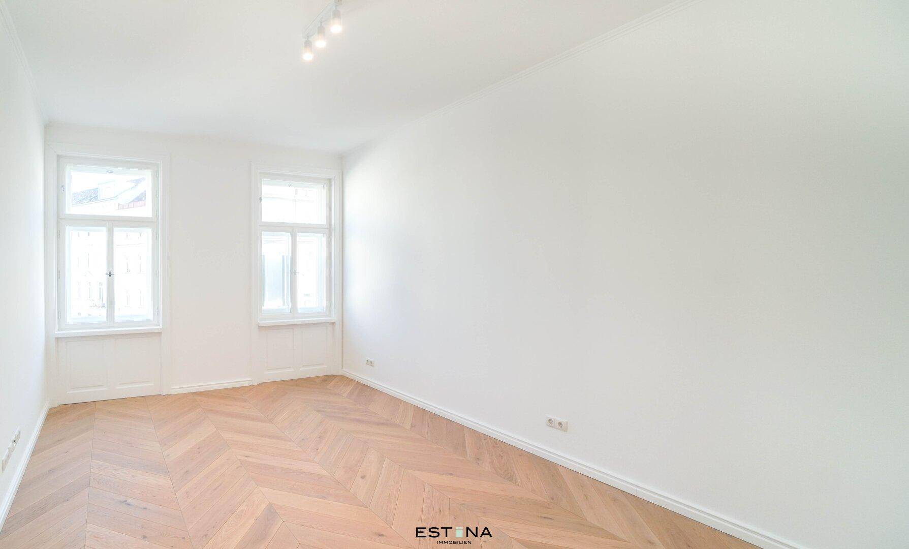 Heller Wohnraum mit zwei Fenstern und elegantem Fischgrätparkett, ideal für vielseitige Nutzung.