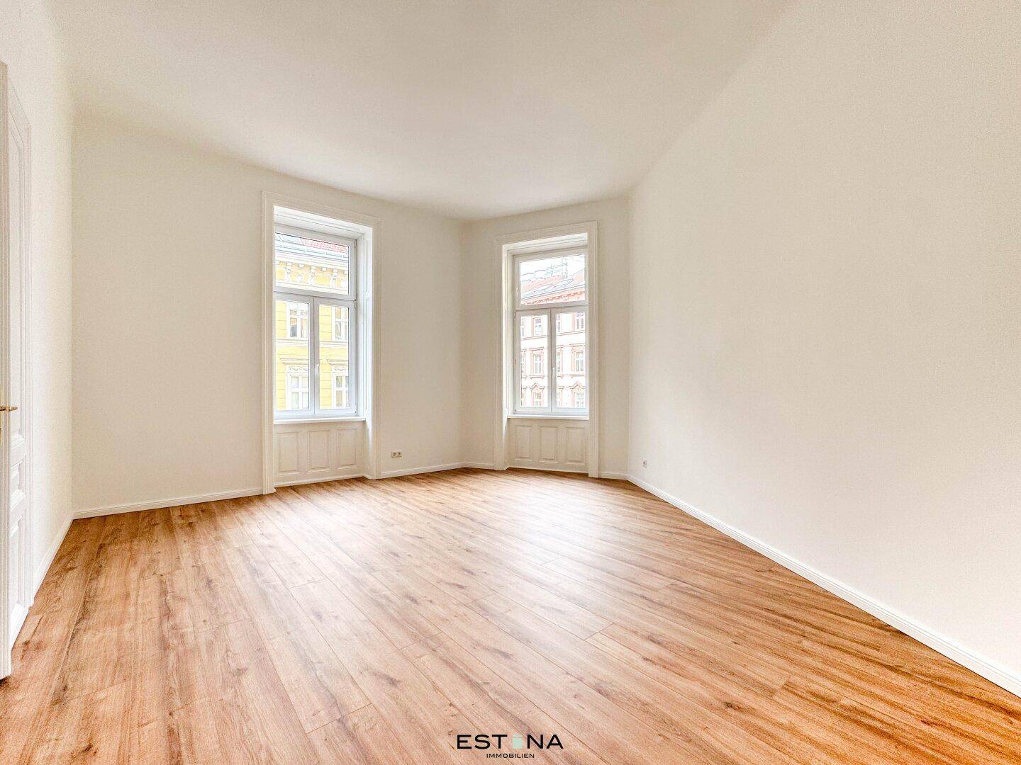 Geräumiges Zimmer mit Holzboden und zwei großen Fenstern, die den Raum erhellen und eine angenehme Atmosphäre schaffen.