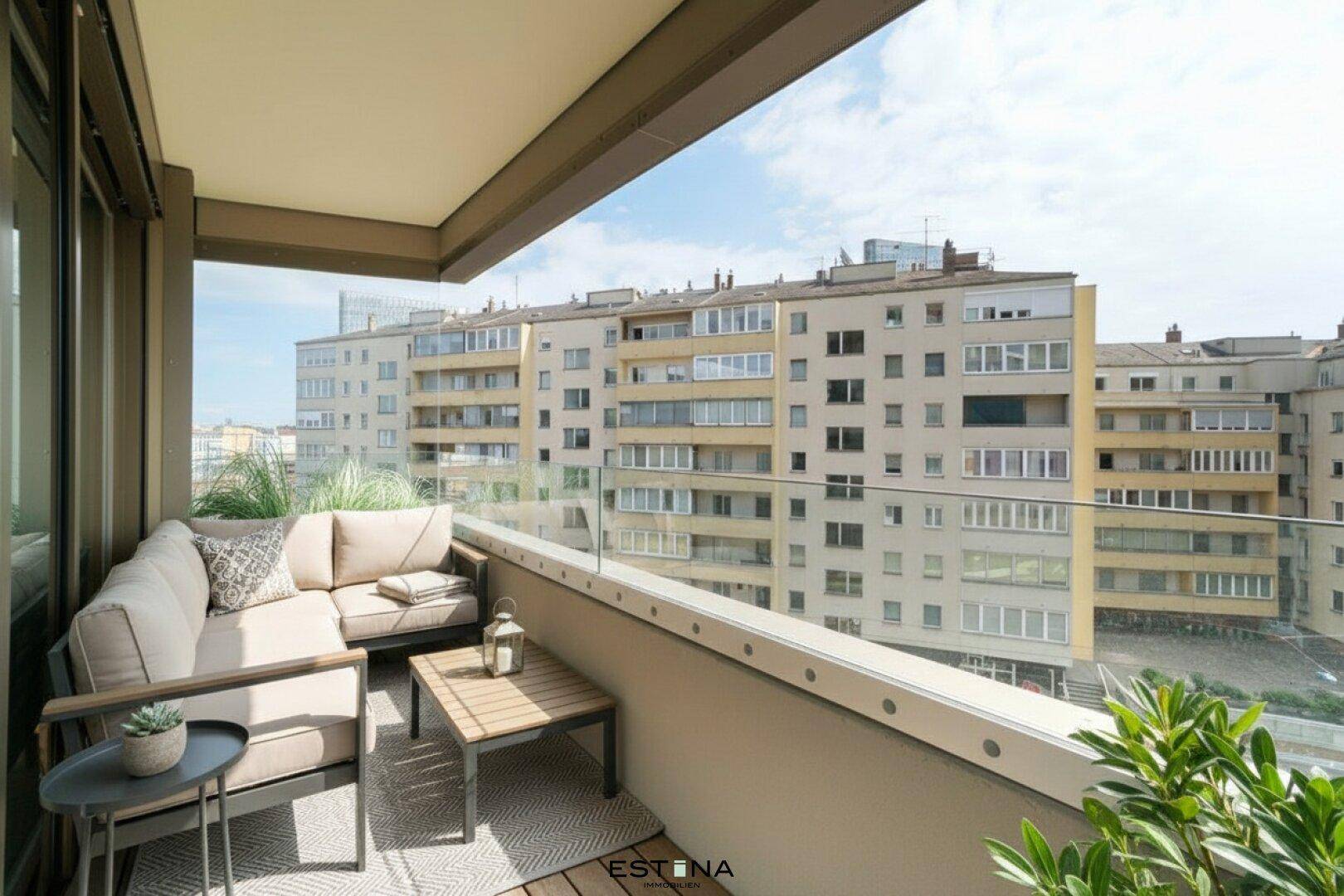 Möblierter Balkon mit Holzboden und Glasgeländer, bietet einen Blick auf die umliegenden Gebäude.