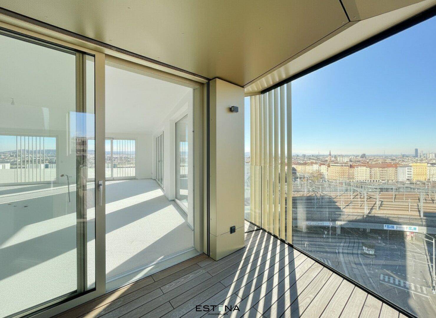 Balkon mit Holzboden und Glasbrüstung, bietet Zugang zu einem hellen Innenraum mit Stadtblick.