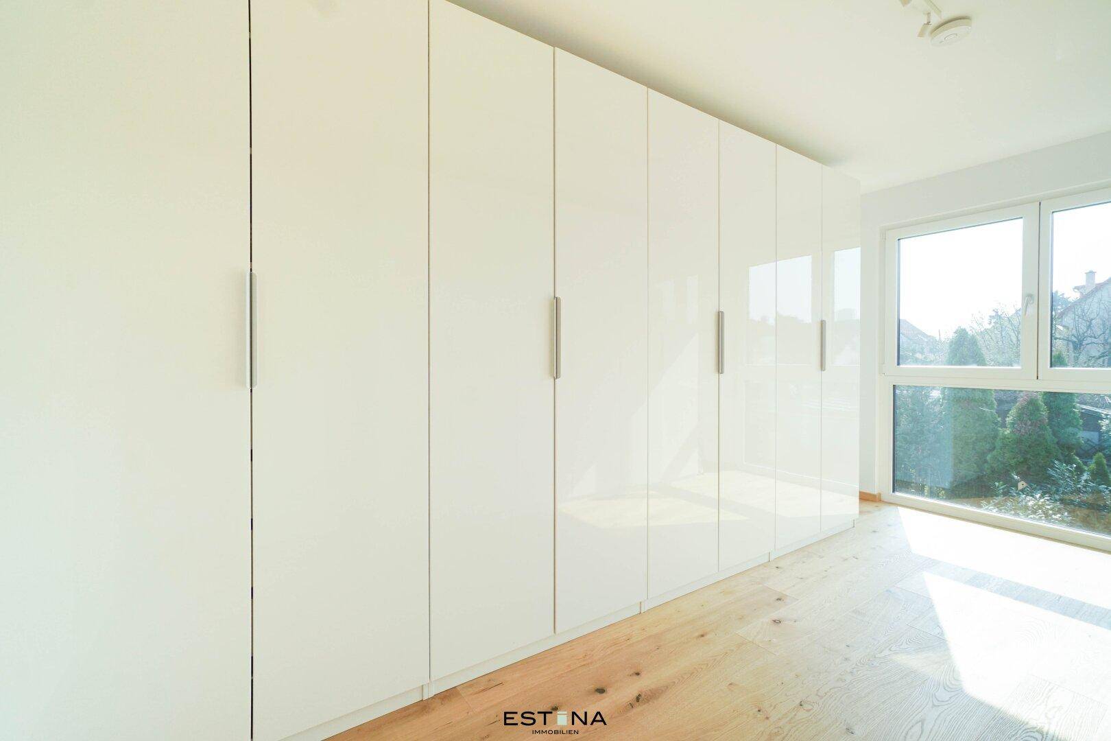 Geräumiges Zimmer mit langen Einbauschränken, Holzboden und großem Fenster mit Grünblick.
