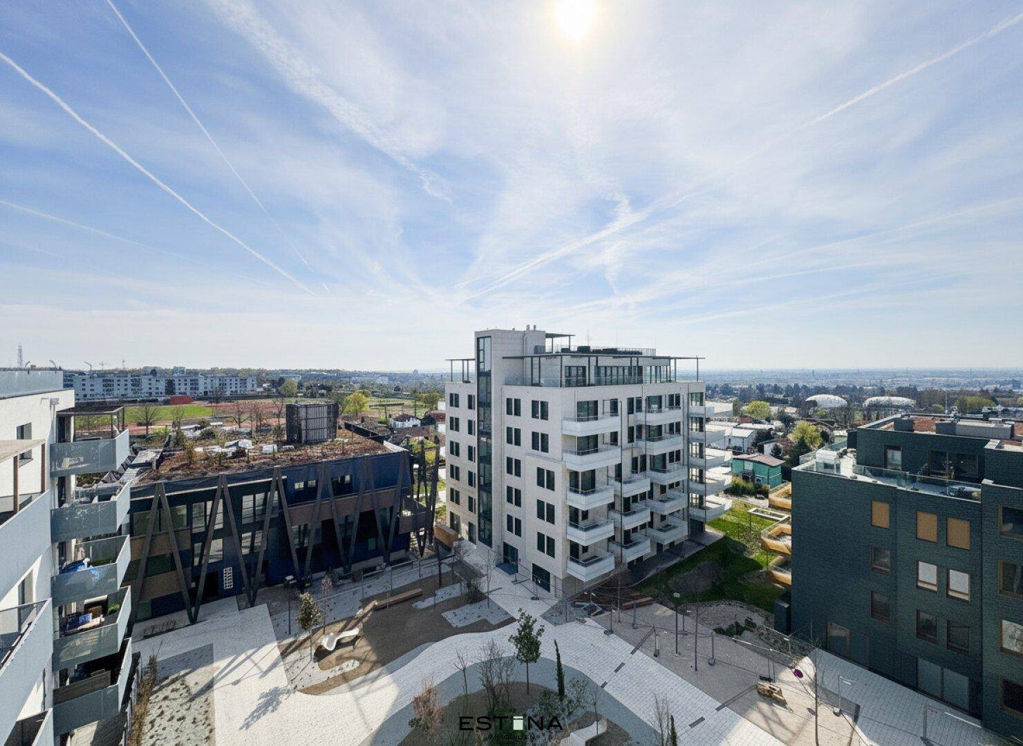 Weitläufige Luftaufnahme eines modernen Wohnkomplexes mit umliegender Stadtlandschaft unter blauem Himmel.