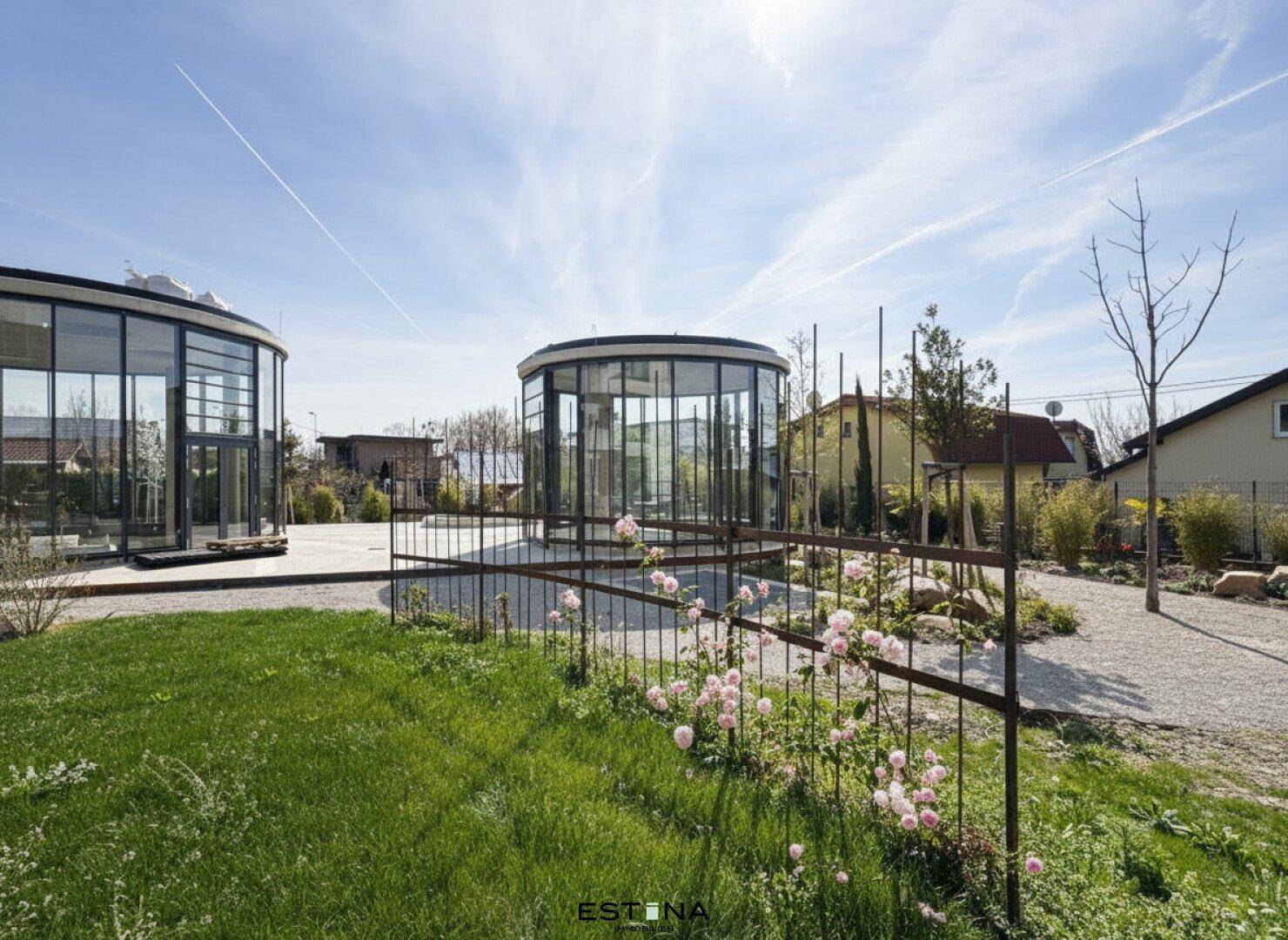 Weitläufiger Gartenbereich mit Rasenflächen, Blumenbeeten und zwei modernen Glaspavillons.