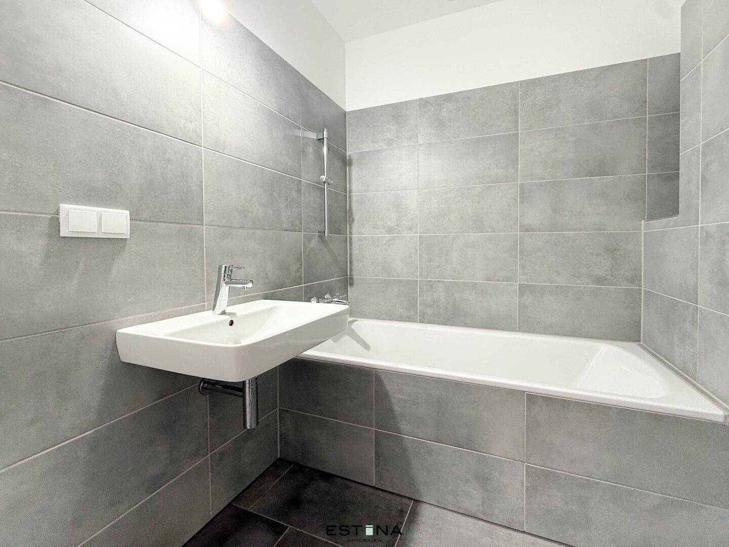 Modernes Badezimmer mit grauen Fliesen, Badewanne und einem einzelnen Waschbecken.