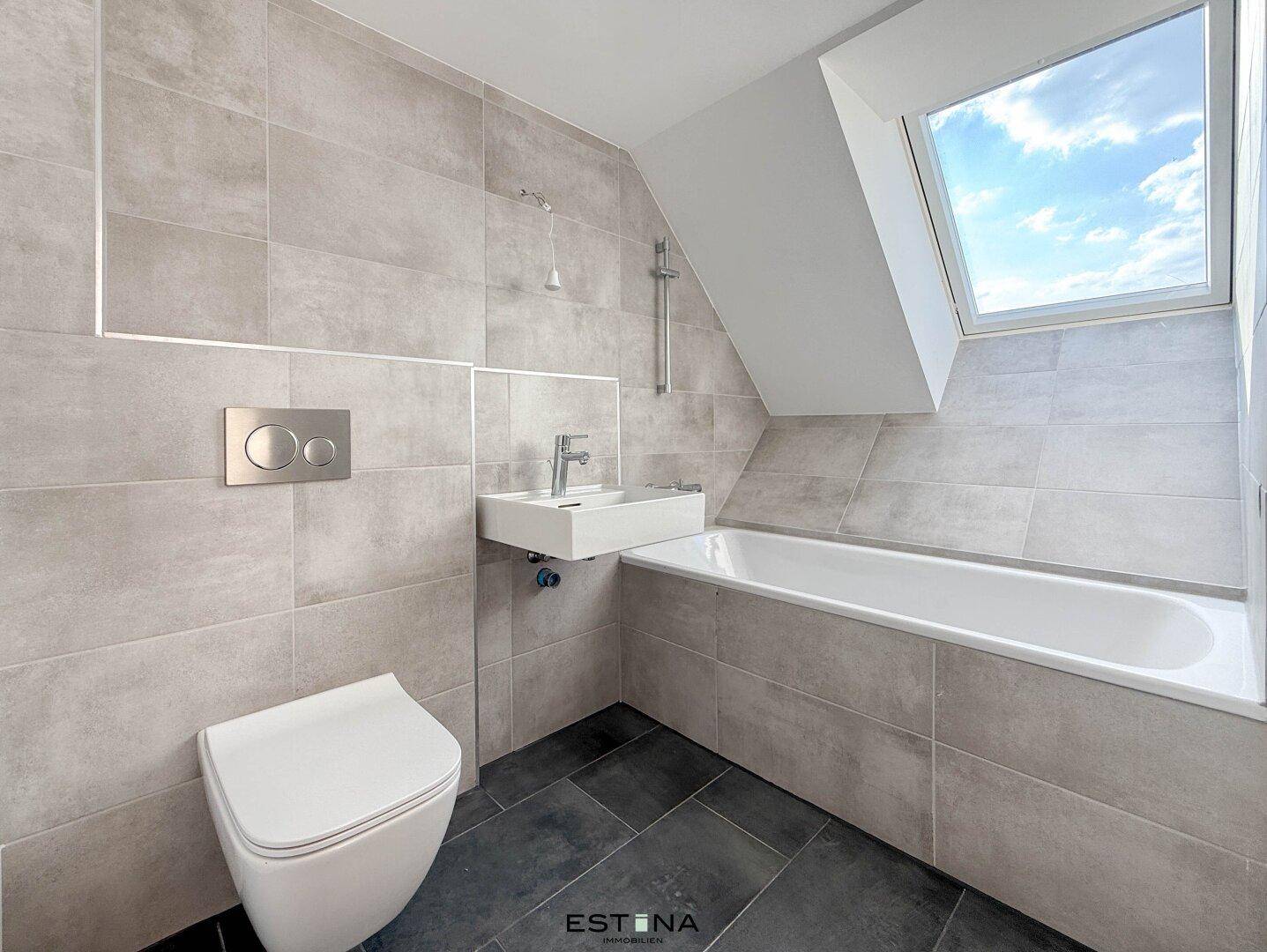 Modernes Badezimmer mit grauen Fliesen, Badewanne, wandhängendem WC und Dachfenster.