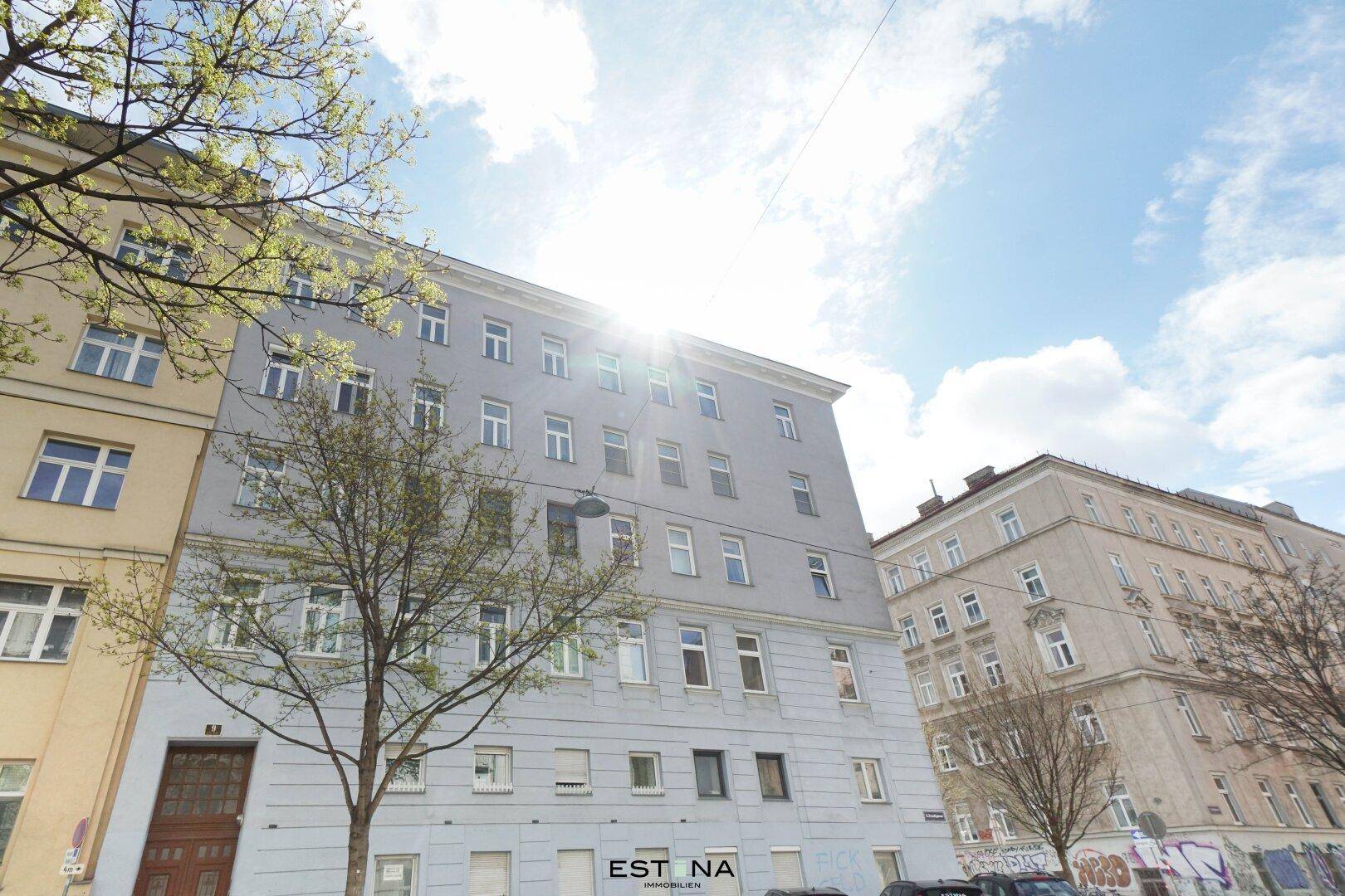 Klassisches Mehrfamilienhaus mit vielen Fenstern und Bäumen davor, bei Sonnenschein.