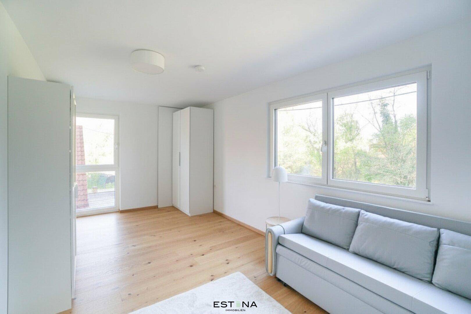 Helles Zimmer mit Schlafsofa, weißen Schränken und Fenstern mit Blick ins Grüne.