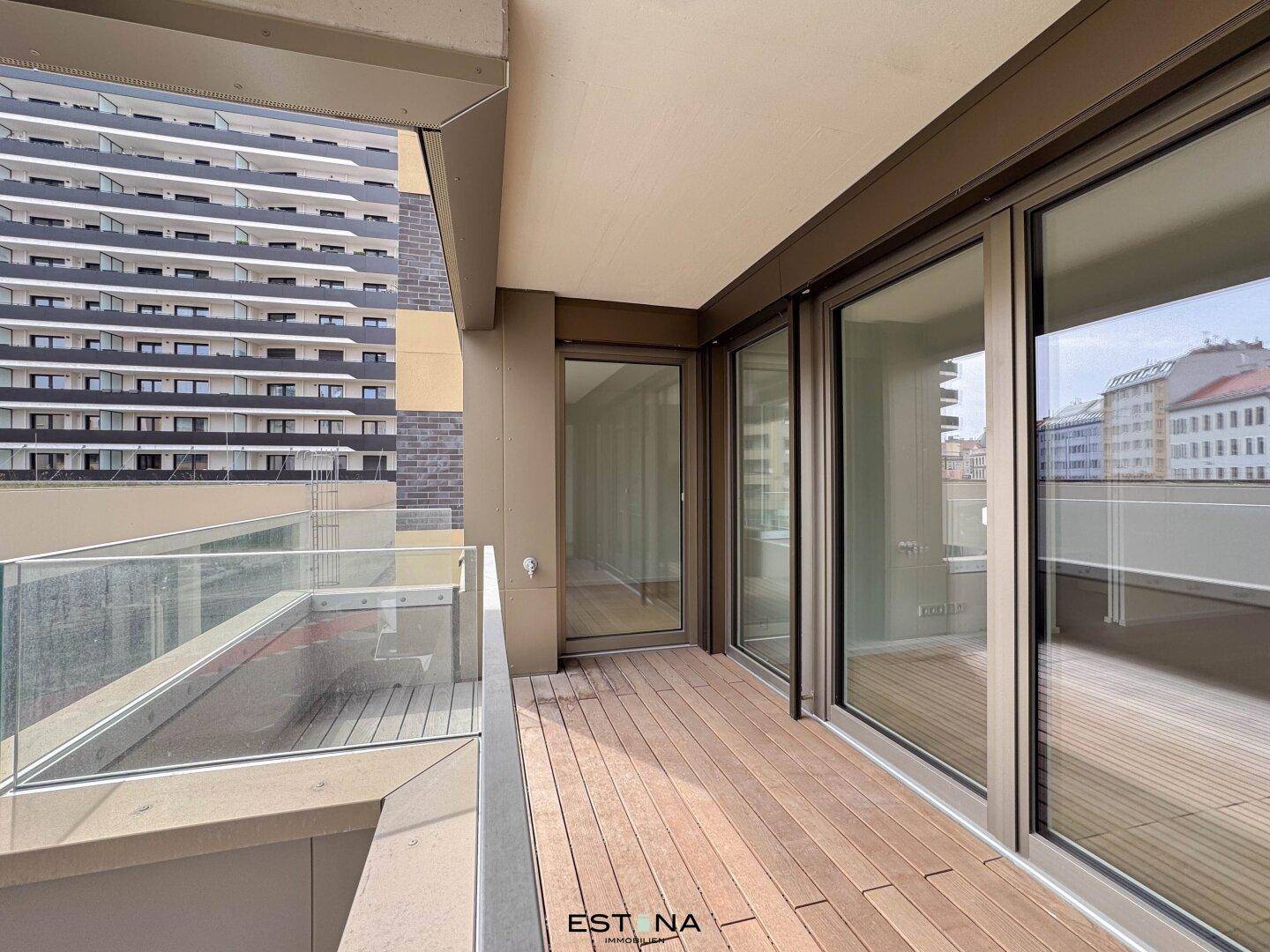 Moderner Balkon mit Holzboden und Glasgeländer, bietet einen Blick auf die umliegenden Gebäude.