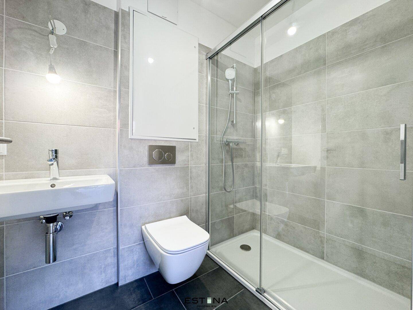 Modernes Badezimmer mit grauen Fliesen, wandhängendem WC und einer begehbaren Dusche.