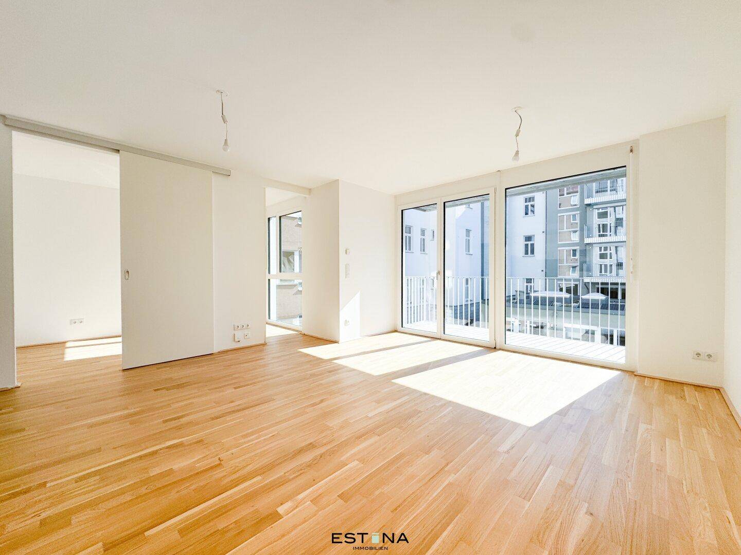 Heller Wohnbereich mit großen Fenstern, Holzboden und Zugang zu einem französischen Balkon.