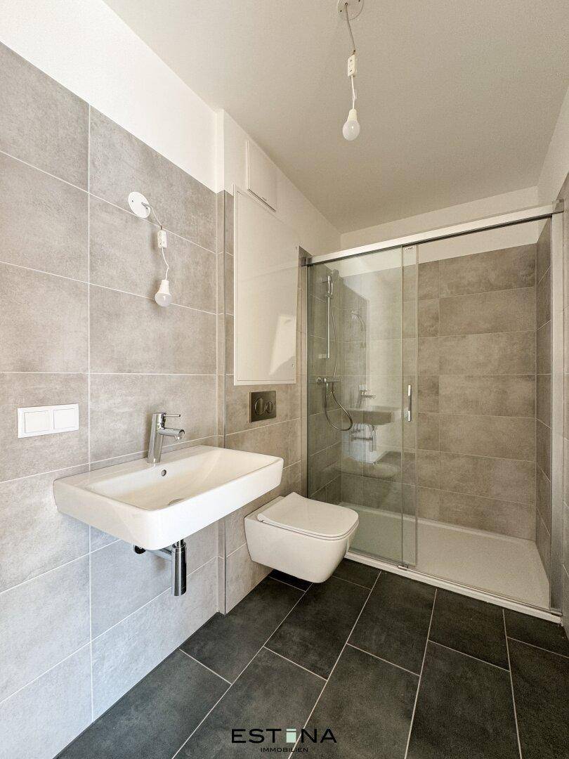 Modernes Badezimmer mit grauen Fliesen, wandhängendem WC und begehbarer Dusche mit Glasabtrennung.