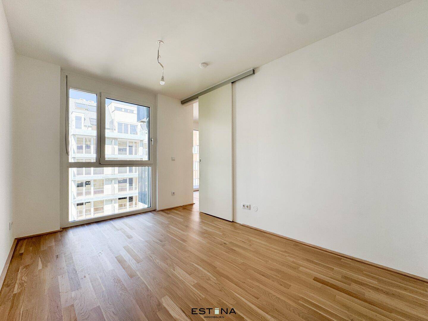 Helles Schlafzimmer mit Parkettboden und großem Fenster, das viel Tageslicht hereinlässt.