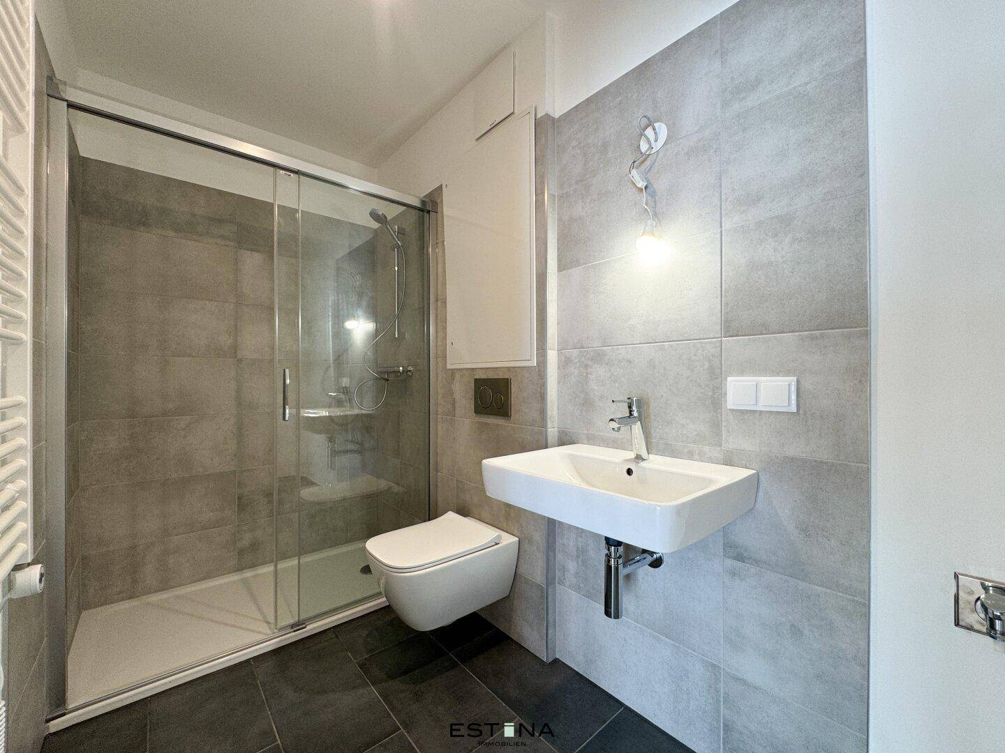 Modernes Badezimmer mit grauen Fliesen, ebenerdiger Dusche, wandhängendem WC und Handtuchheizkörper.