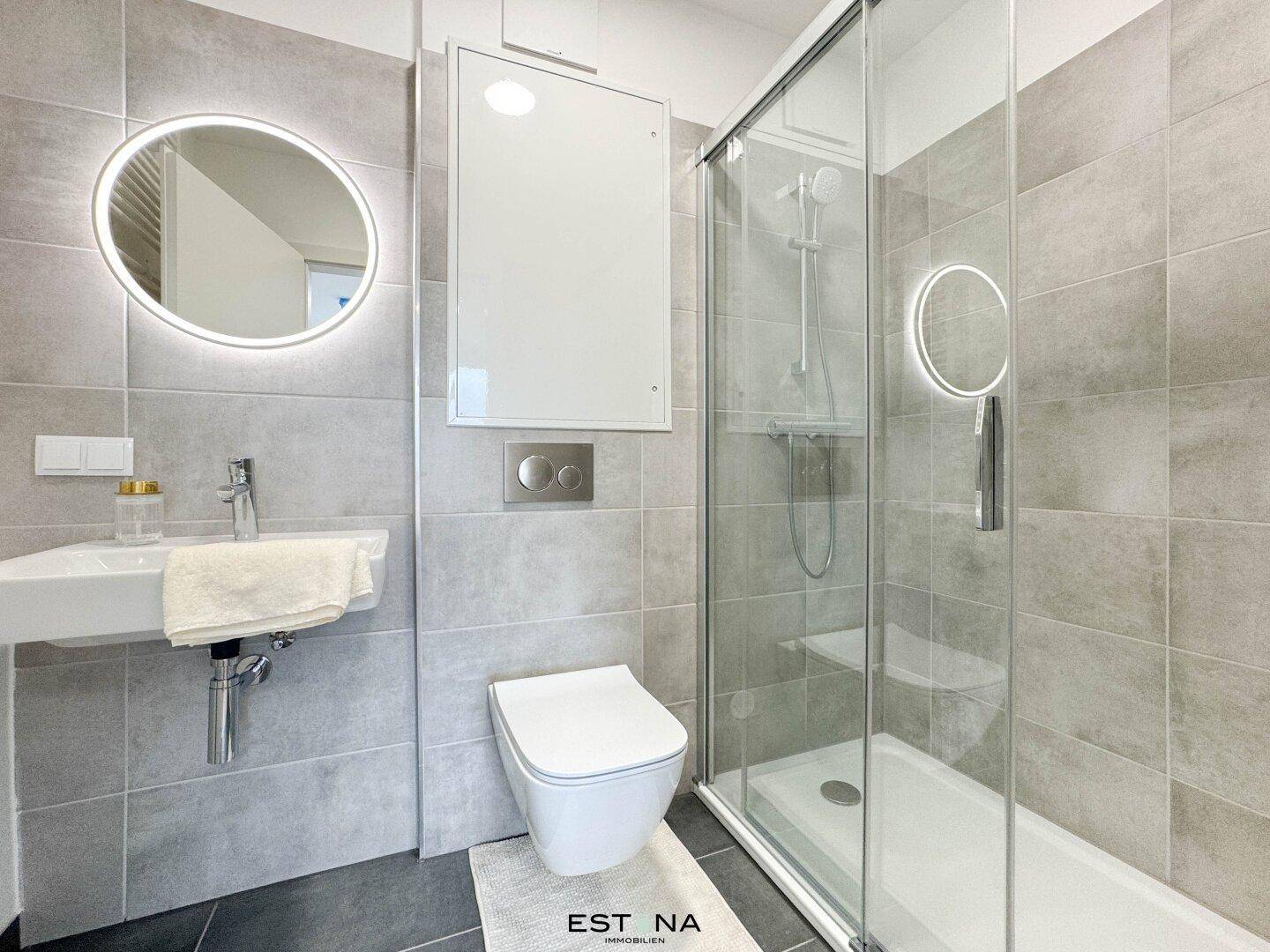 Modernes Badezimmer mit grauen Fliesen, begehbarer Dusche und beleuchtetem Spiegel.