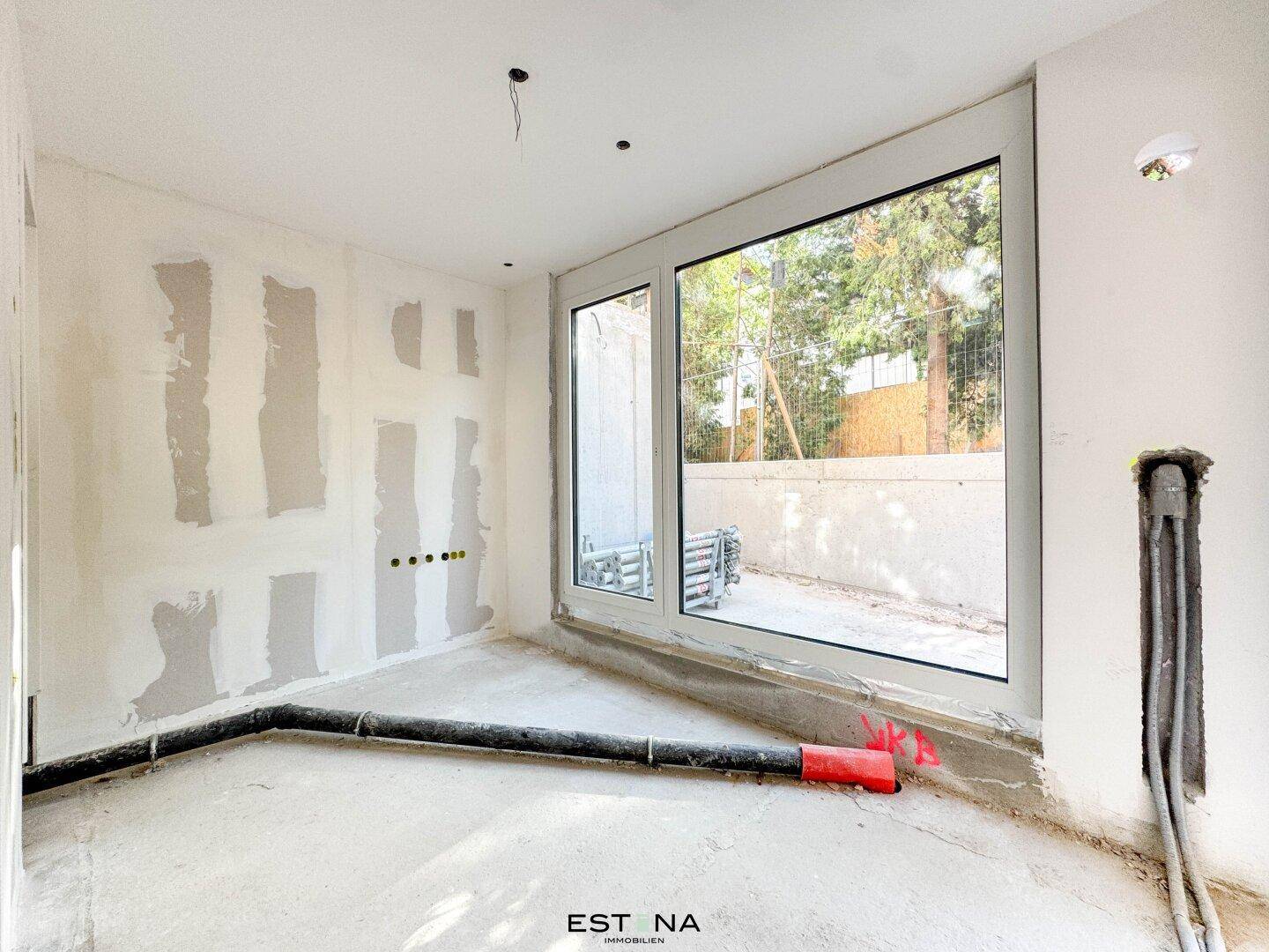 Rohbauzimmer mit großem Fenster, vorbereiteten Wänden und sichtbaren Leitungen für zukünftige Installationen.