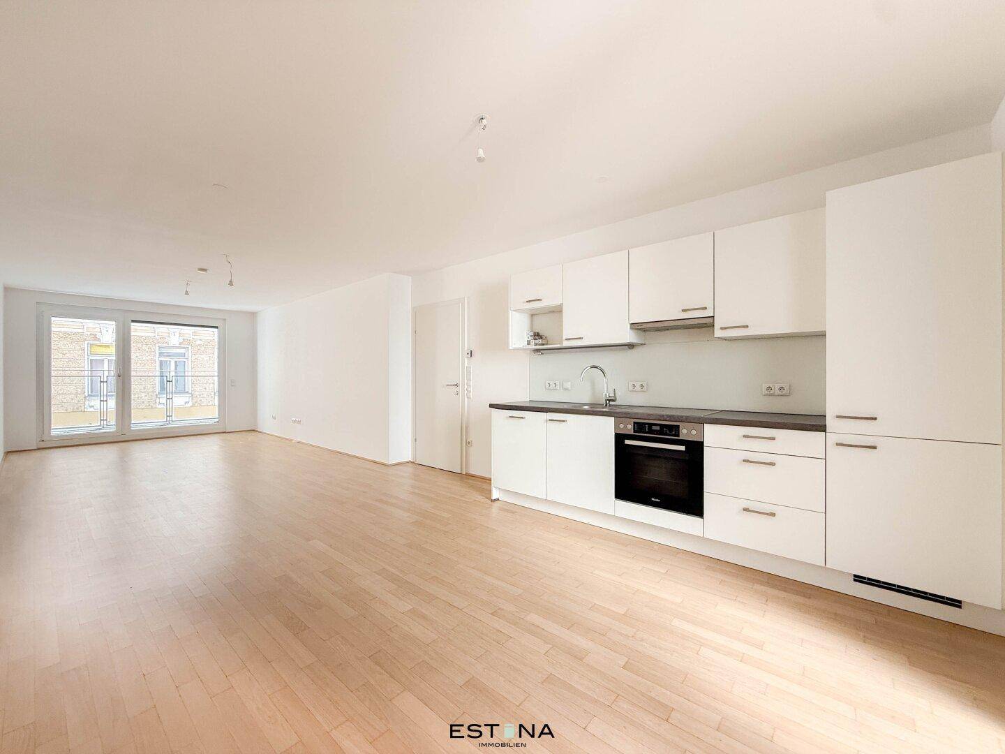 Offener Wohnbereich mit integrierter moderner Küche, hellem Parkettboden und Zugang zum Balkon.