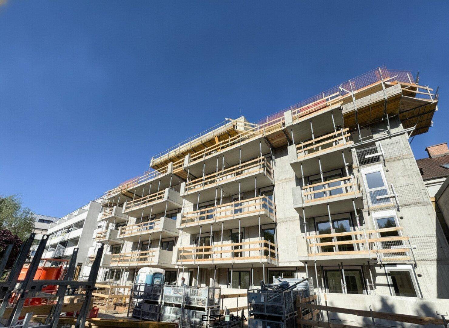 Neubau im Bauzustand mit sichtbaren Betonstrukturen, Balkonen und Gerüsten unter blauem Himmel.