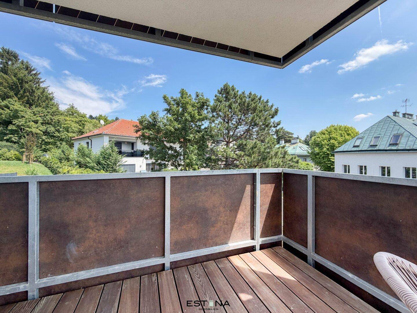 Sonniger Balkon mit Holzboden und Blick ins Grüne, umgeben von Bäumen und Wohnhäusern.