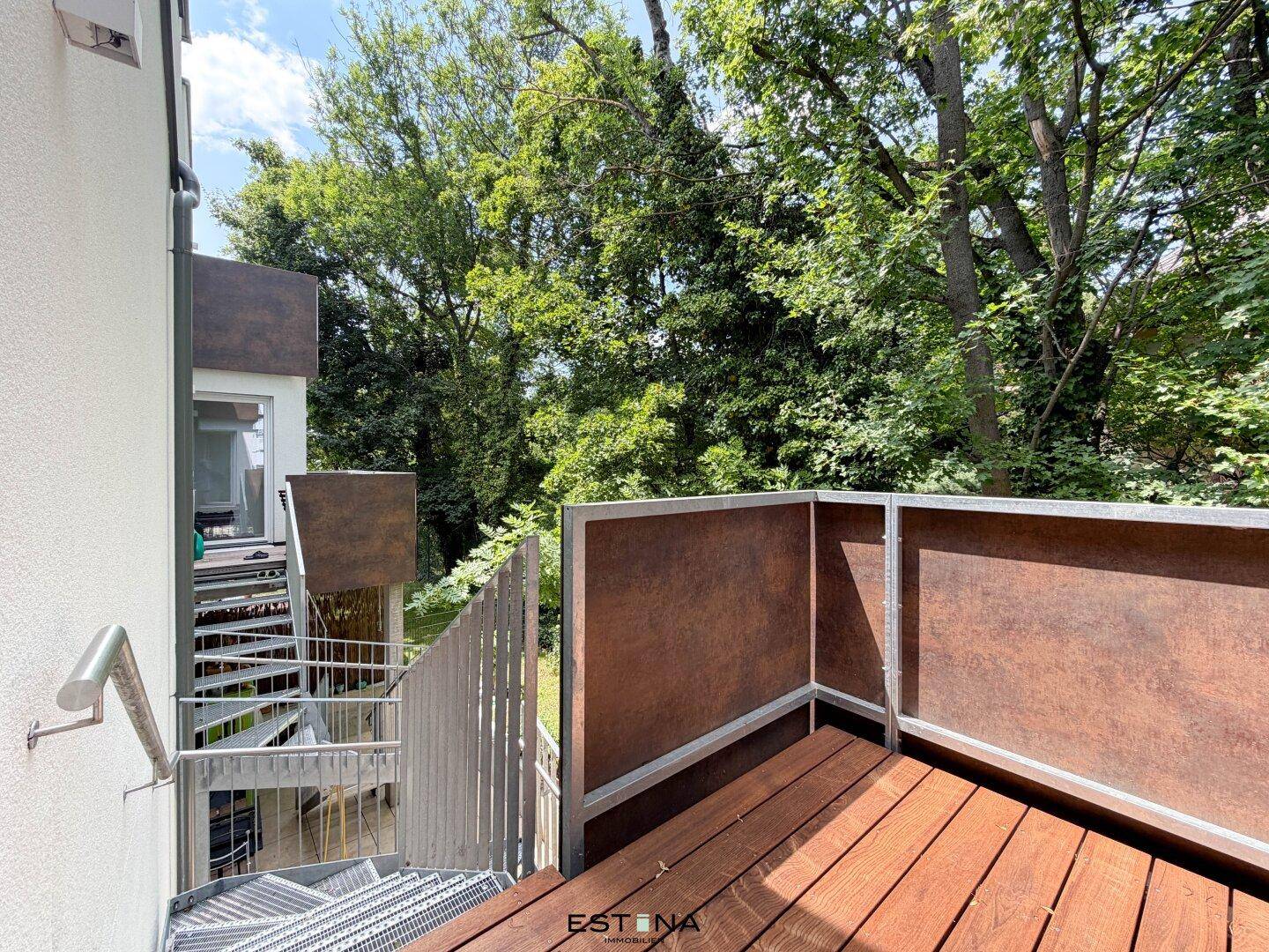 Balkon mit Holzboden und Außentreppe, umgeben von üppigem Grün und Bäumen.