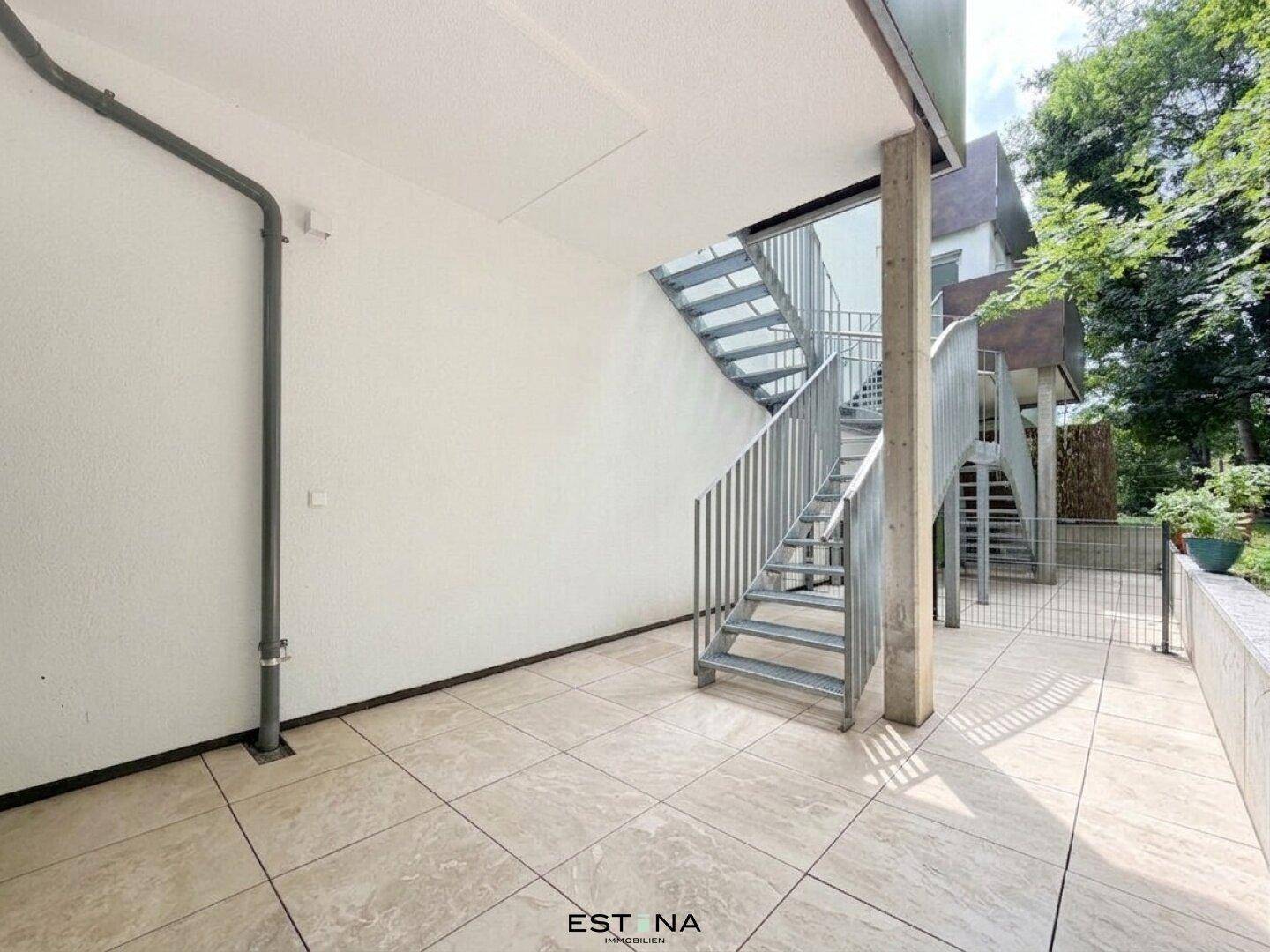 Große geflieste Terrasse mit Außentreppe und Zugang zum Gartenbereich, umgeben von Bäumen.