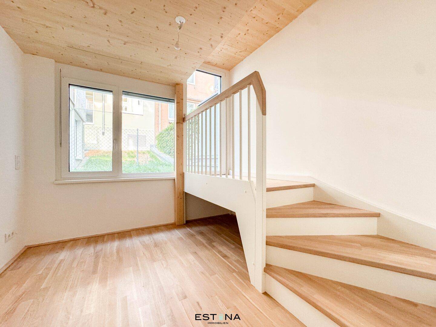 Heller Wohnbereich mit Holztreppe, Parkettboden und großen Fenstern, die viel Tageslicht hereinlassen.