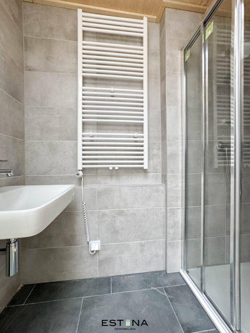 Modernes Badezimmer mit grauen Fliesen, einer Duschkabine und einem praktischen Handtuchheizkörper.