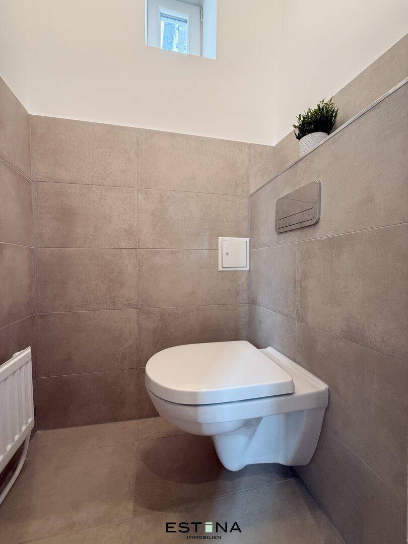 Separates WC mit wandhängender Toilette und kleinem Fenster, in modernen Fliesen ausgeführt.