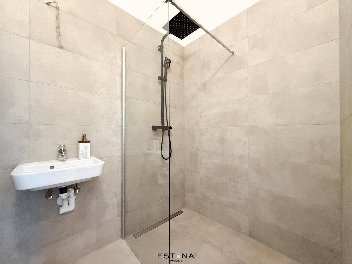 Modernes Badezimmer mit ebenerdiger Dusche und kleinem Waschbecken, in hellen Fliesen gehalten.