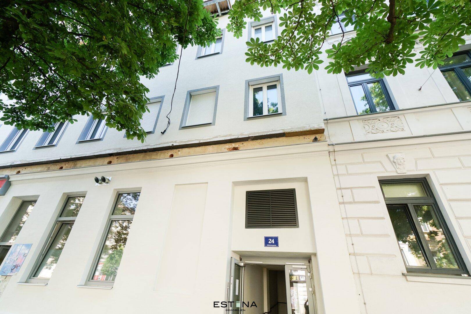 Fassade eines Mehrfamilienhauses mit modernen Fenstern und Eingangsbereich unter Bäumen.