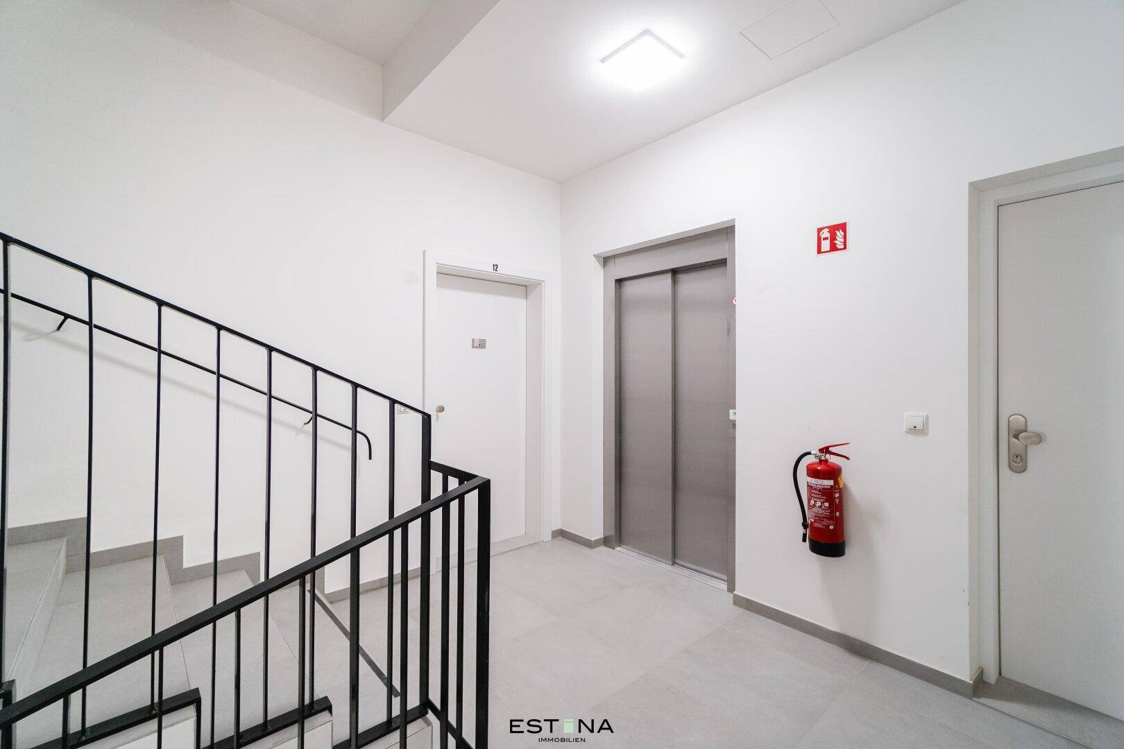 Treppenhaus und Flur mit Aufzug und Brandschutzvorrichtungen, gut beleuchtet und gepflegt.