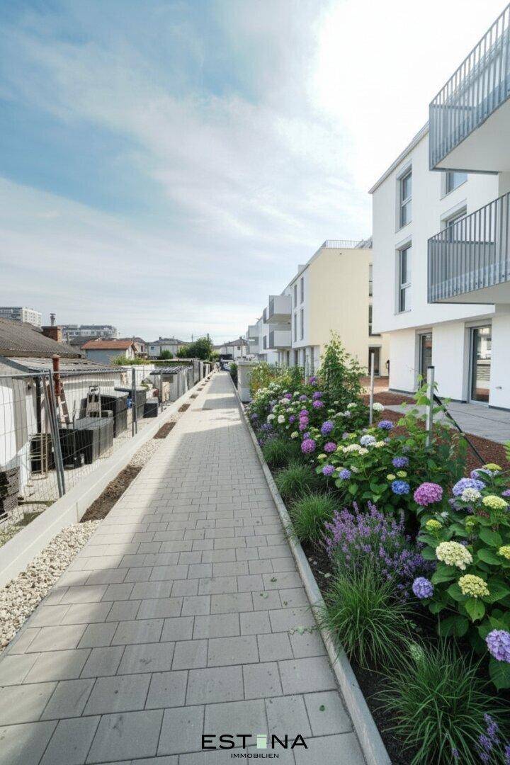 Gepflasterter Gehweg entlang einer modernen Wohnanlage mit blühenden Blumenbeeten.