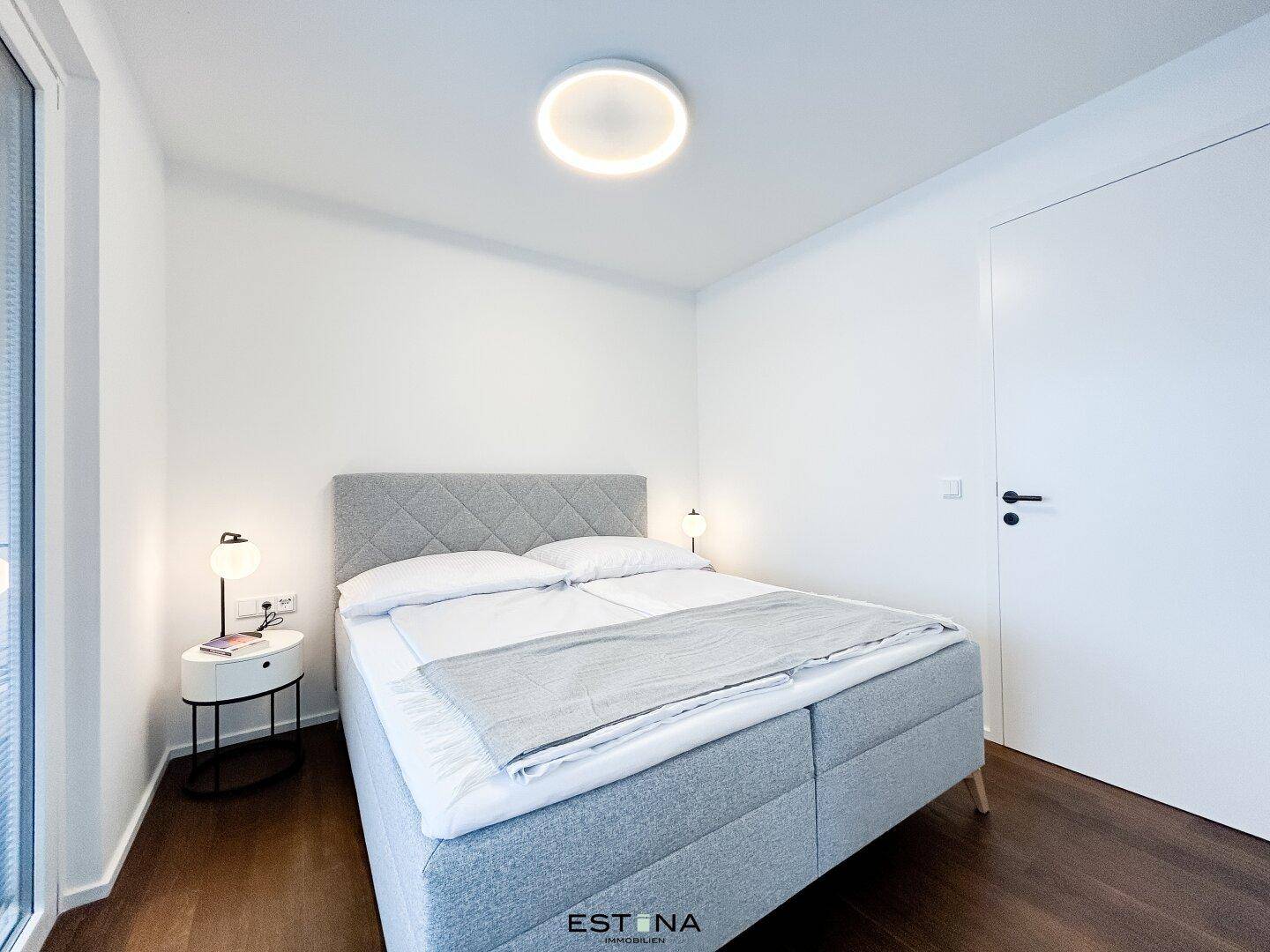 Modernes Schlafzimmer mit grauem Polsterbett, Holzboden und schlichter, heller Einrichtung.
