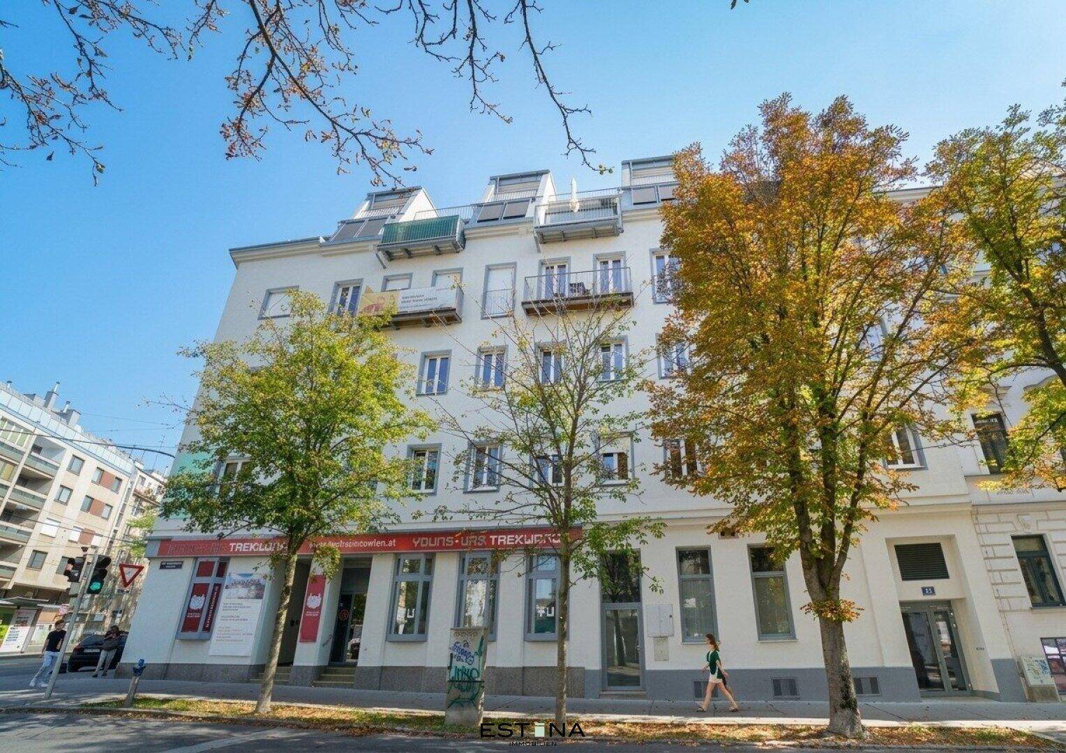 Gesamtansicht eines modernen Mehrfamilienhauses mit Balkonen, Geschäften im Erdgeschoss und Bäumen.