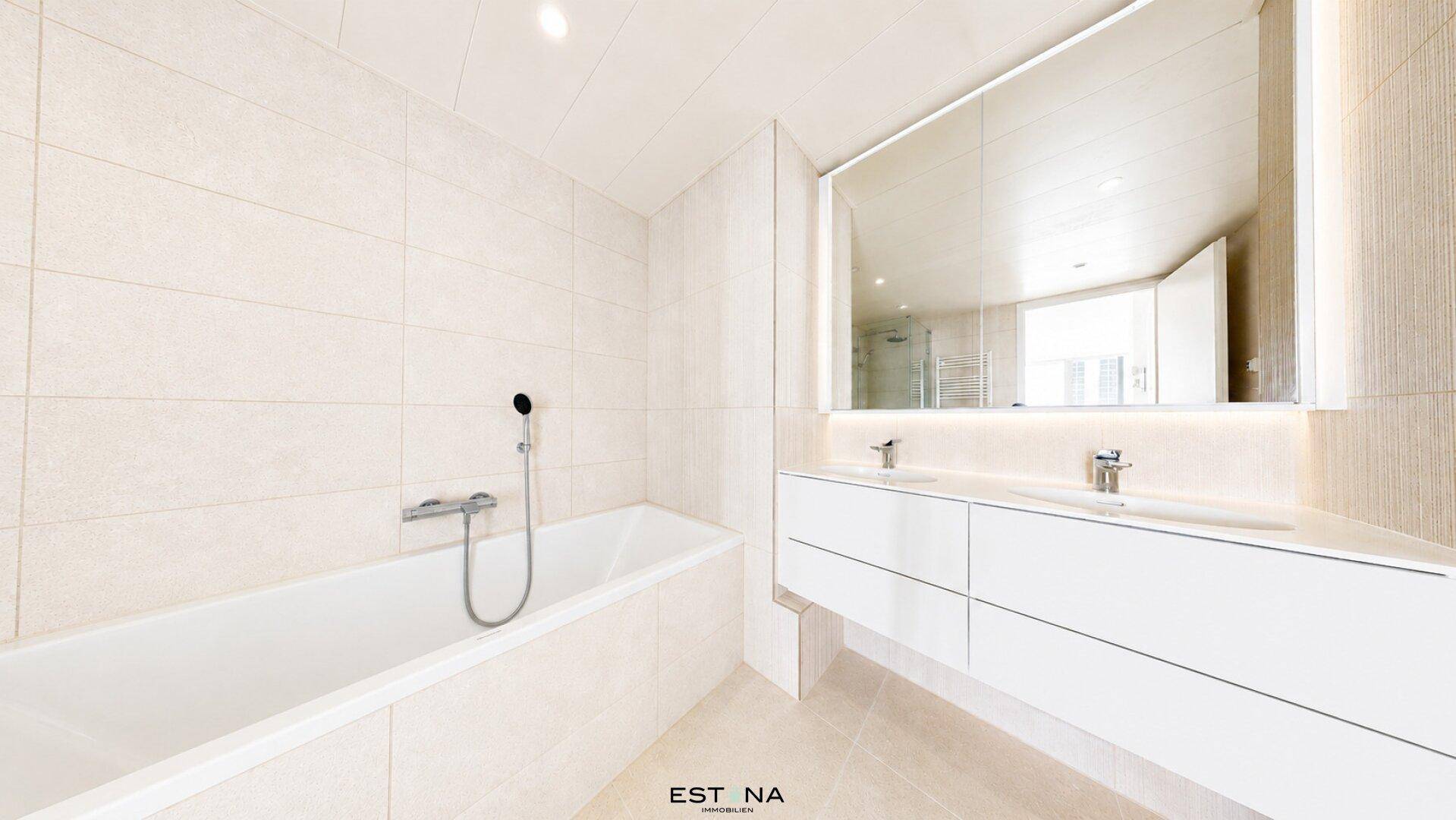 Modernes Badezimmer mit heller Fliesenoptik, Badewanne und Doppelwaschbecken mit beleuchtetem Spiegel.