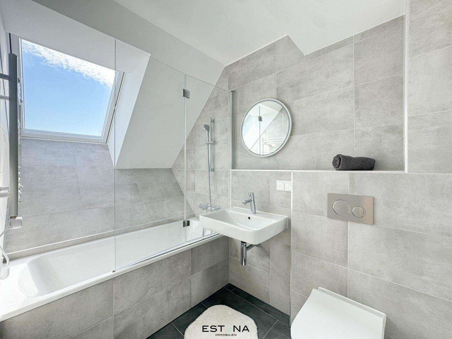 Modernes Badezimmer mit grauen Fliesen, Badewanne und Dachfenster für natürliches Licht.