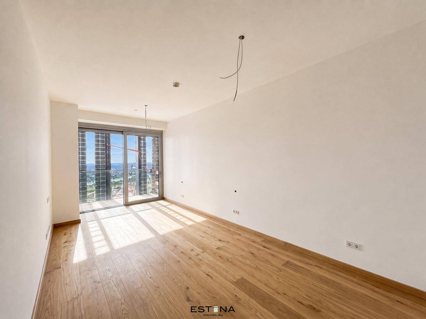 Leeres Schlafzimmer mit Holzboden und großen Fenstern, die viel Tageslicht hereinlassen.