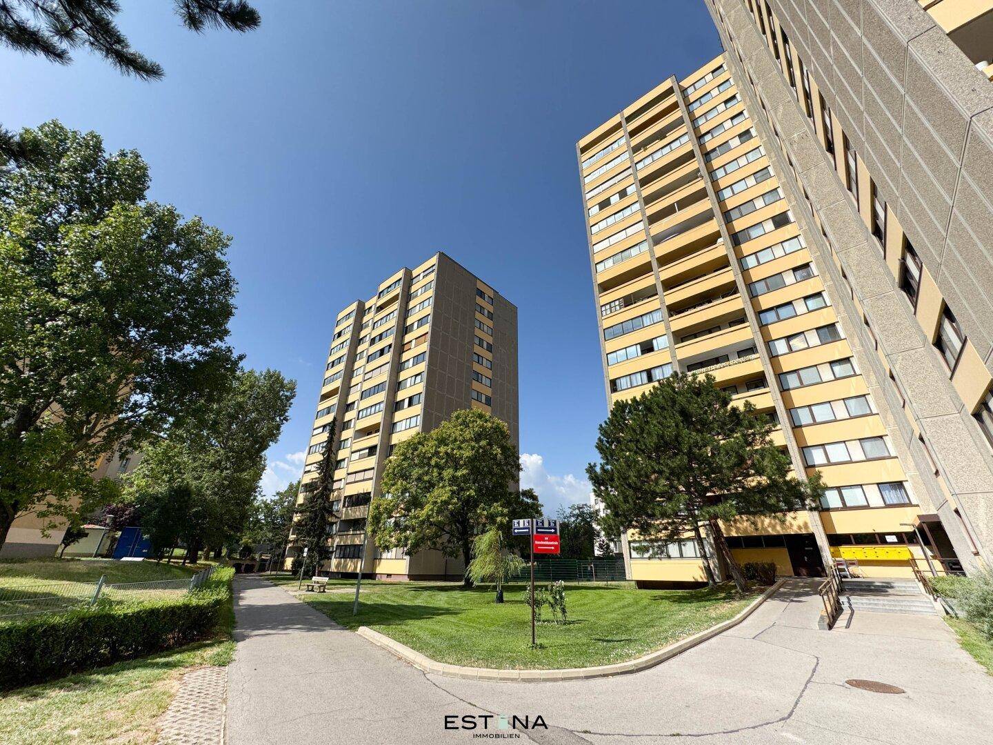 Zwei hohe Wohngebäude in einer grünen Umgebung mit Bäumen und Wegen, viel Freifläche.
