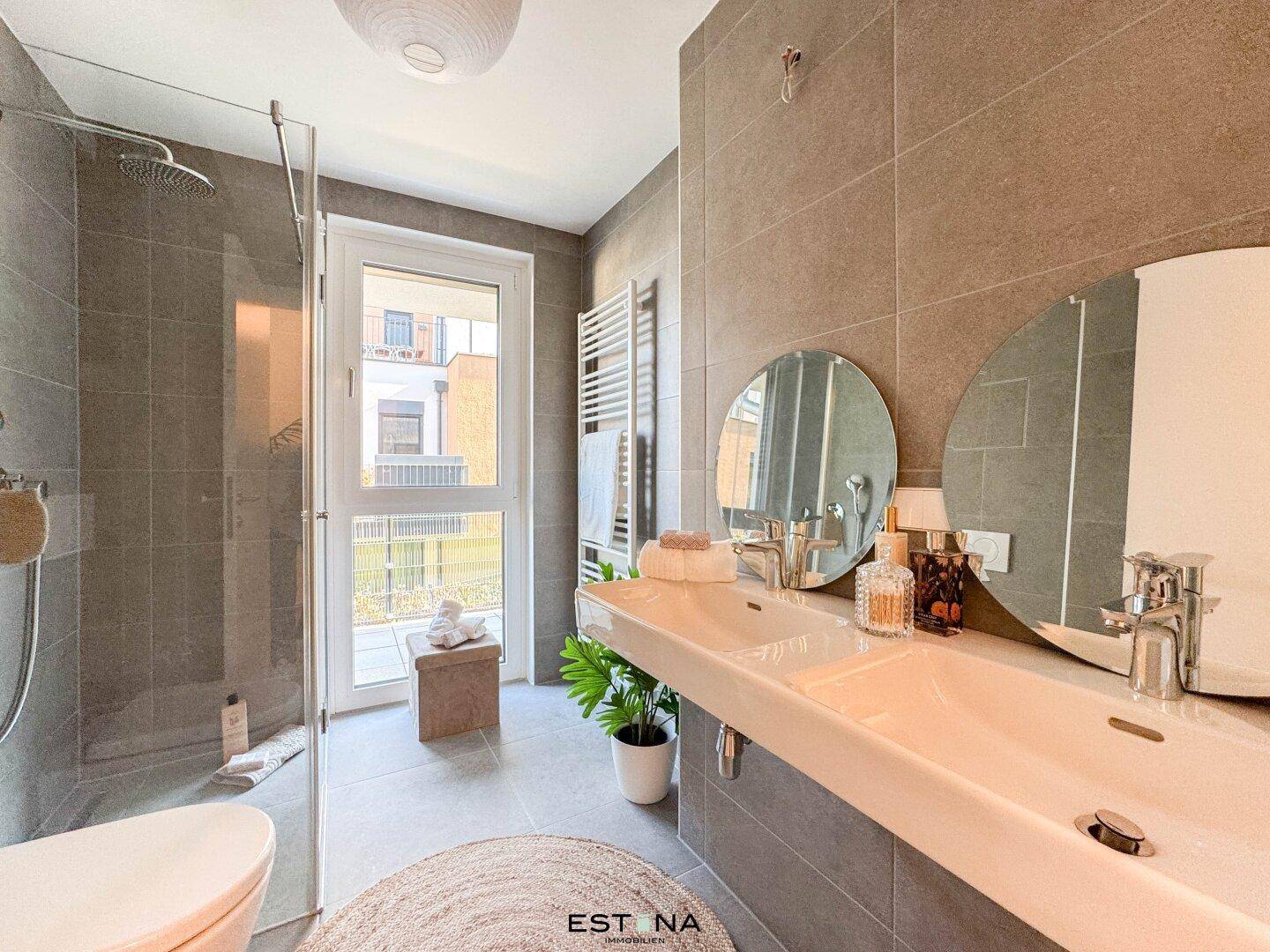 Modernes Badezimmer mit grauen Fliesen, Doppelwaschbecken, ebenerdiger Dusche und Fenster für Tageslicht.
