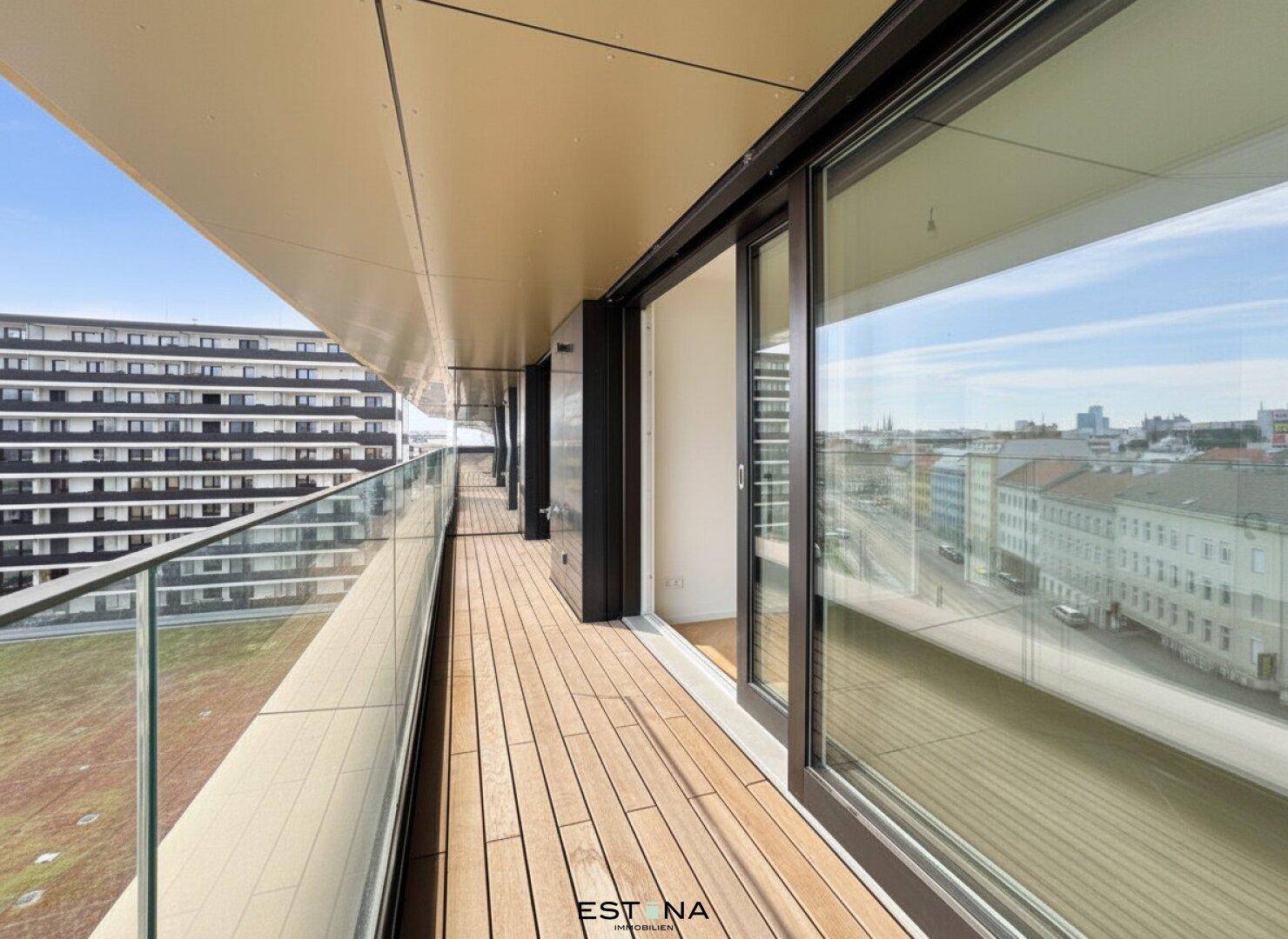 Langer Balkon mit Holzbelag und Glasgeländer, bietet einen weiten Blick über die städtische Umgebung.