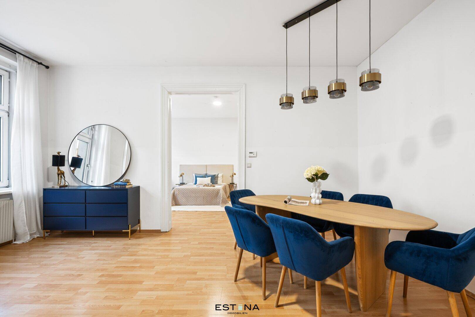 Heller Essbereich mit Holztisch, blauen Stühlen und Blick in das angrenzende Schlafzimmer.
