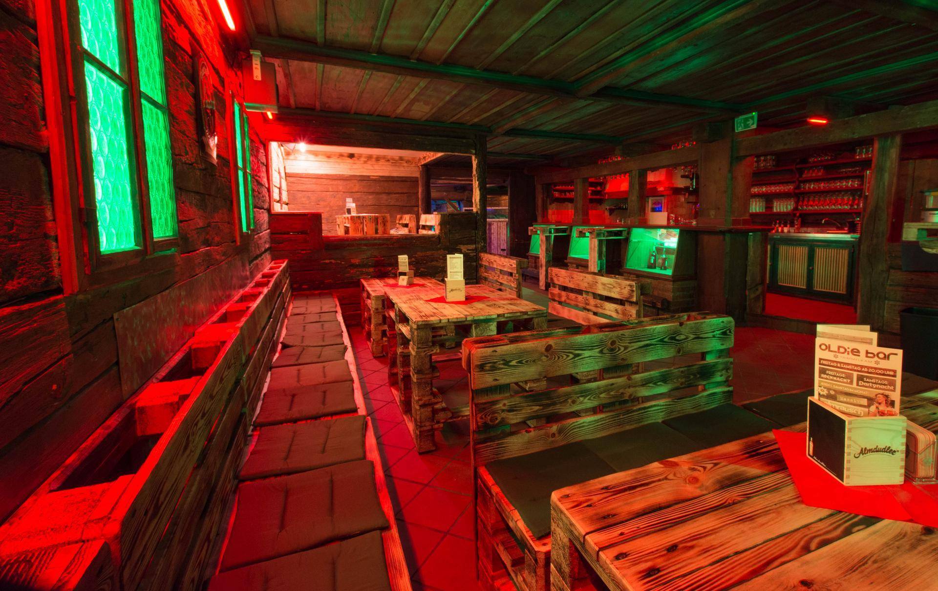 Gemütlicher Sitzbereich mit Palettenmöbeln, Holztischen und grünen Fenstern in roter Lichtatmosphäre.