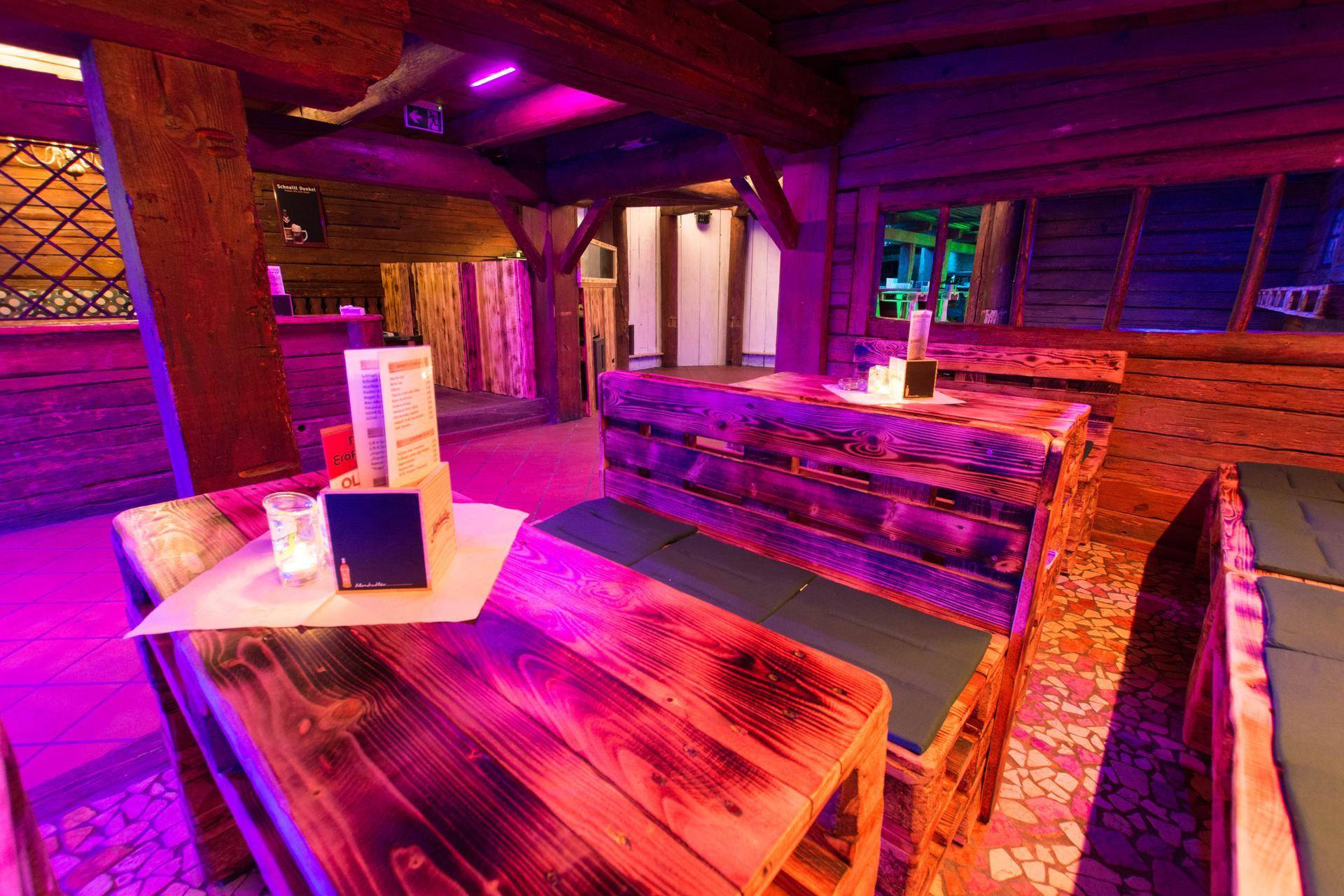 Gemütlicher Sitzbereich mit Palettenmöbeln und Holztischen unter violetter Beleuchtung.